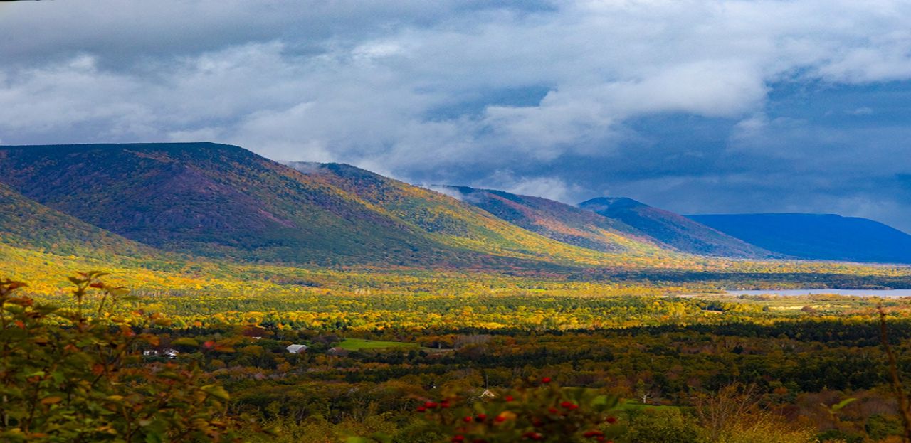 Aerial view of a Nova Scotia valley in peak autumn colour, with forested hillsides displaying yellow, orange, red, and green foliage   beneath a dramatic cloudy sky, small farm buildings visible in the valley below.